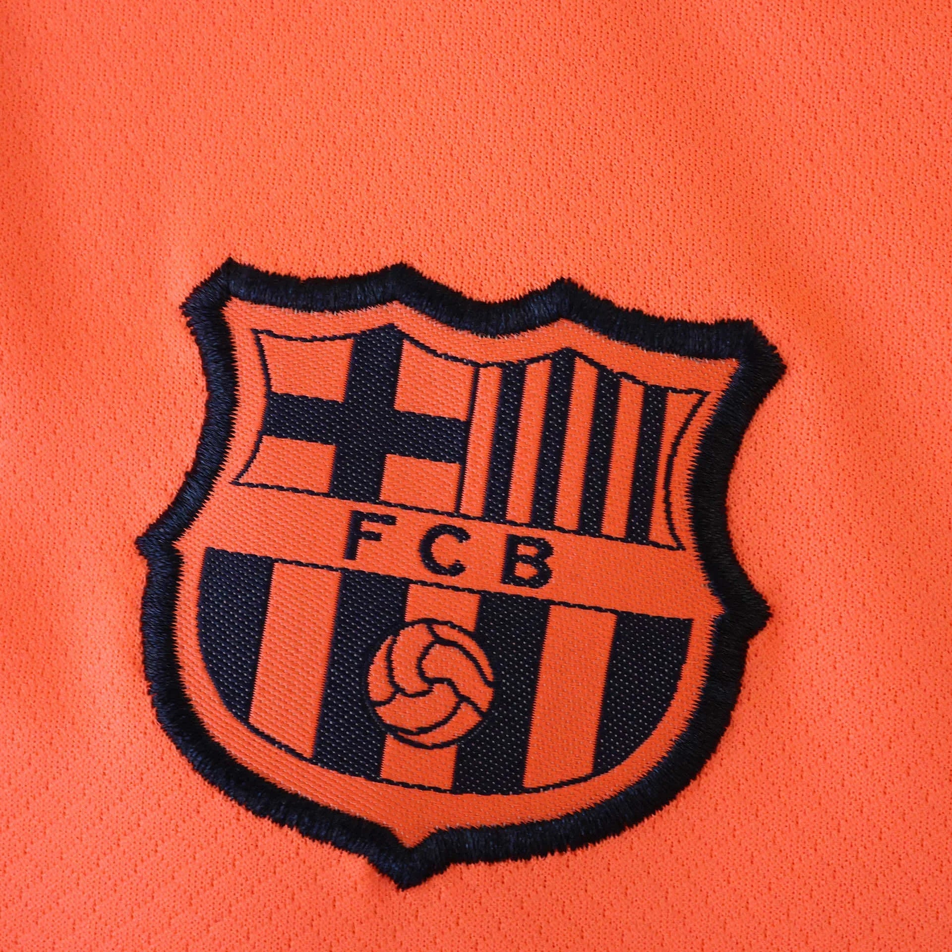 Detalhe do escudo bordado na camisa infantil oficial do FC Barcelona. Parte do kit Mini Mantos laranja