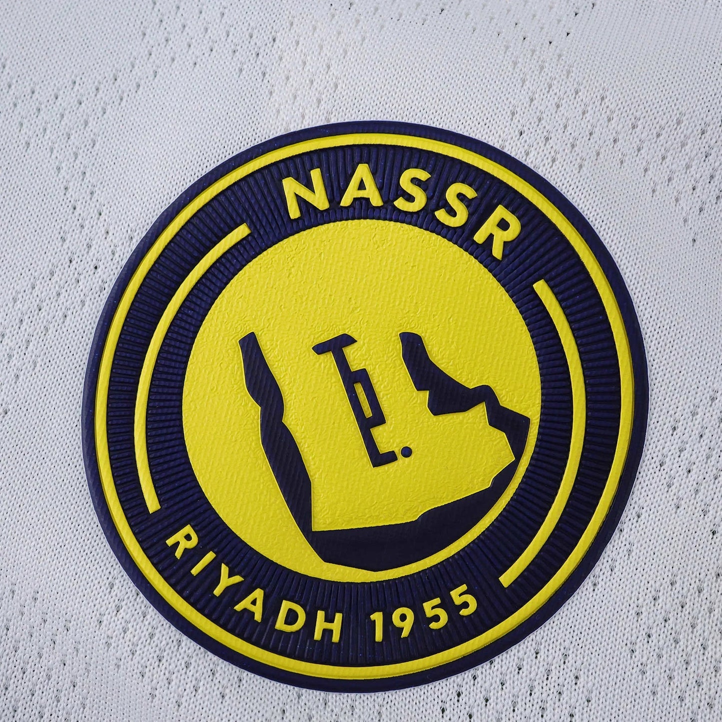 Escudo do Al Nassr em relevo azul e amarelo sobre tecido branco texturizado de camisa de futebol infantil.