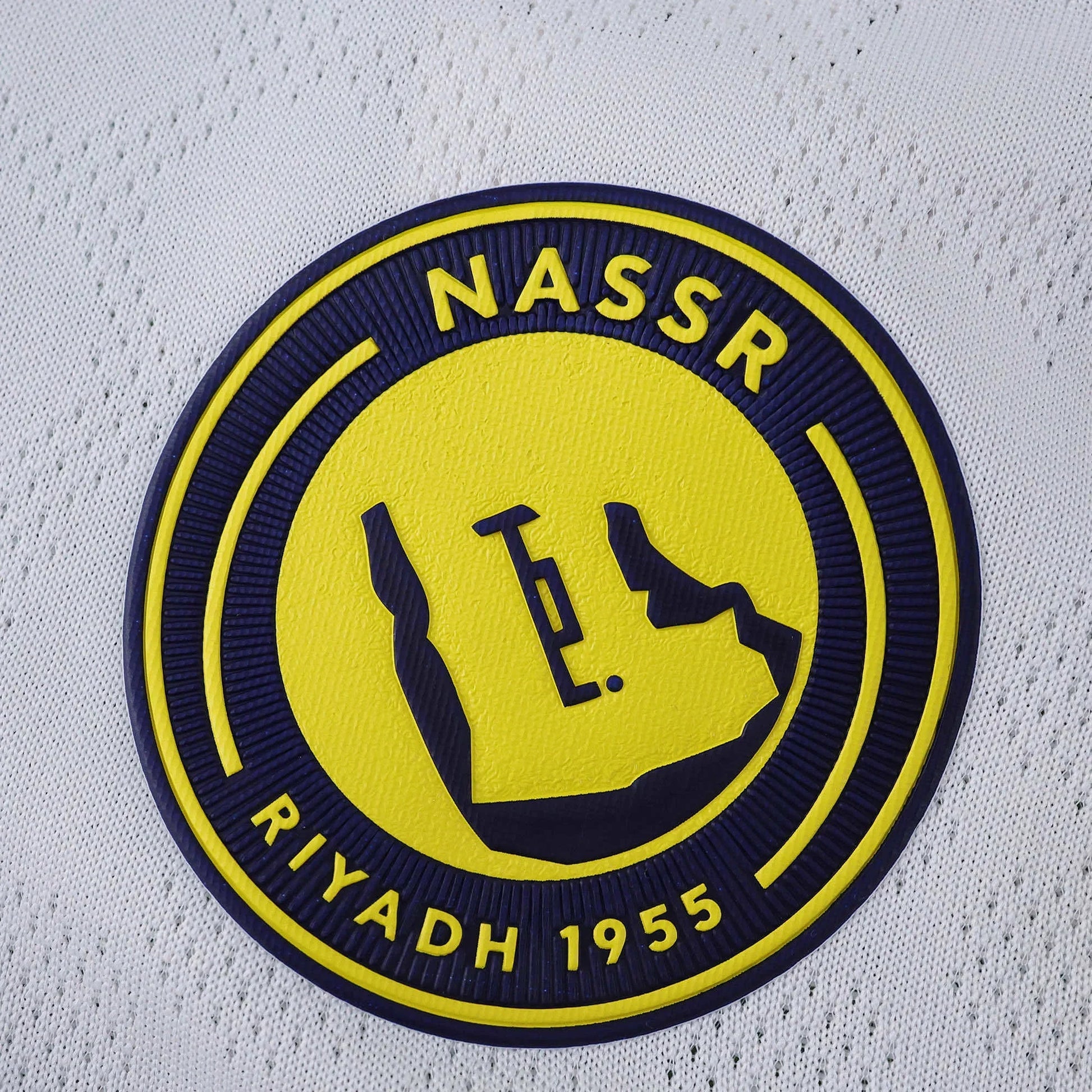 Escudo do Al Nassr em relevo azul e amarelo sobre tecido branco texturizado de camisa de futebol infantil.