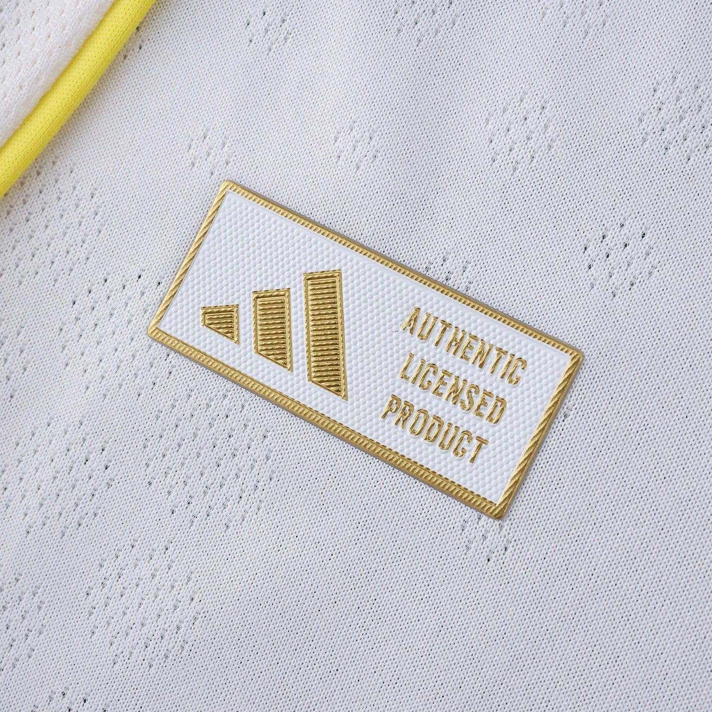 Close-up da camisa branca de futebol com um selo retangular branco e dourado escrito 'Authentic Licensed Product'.