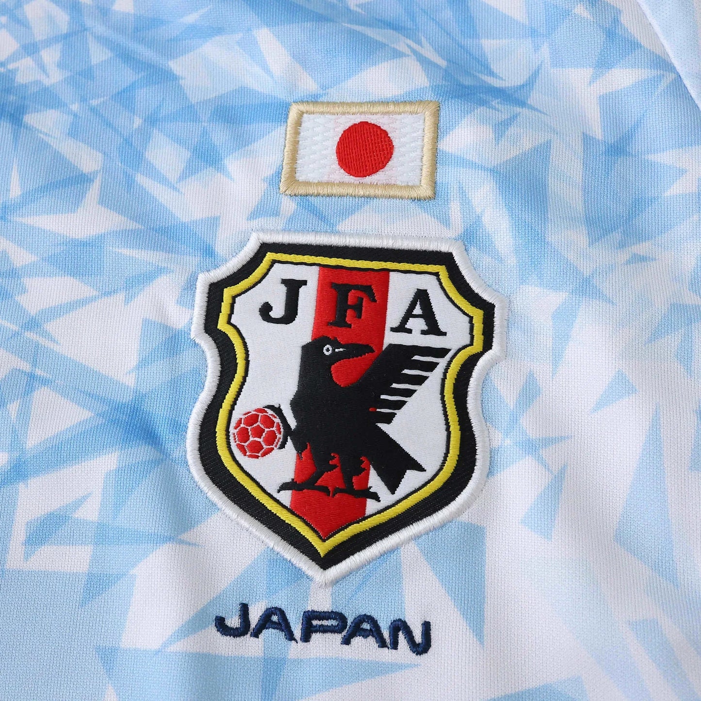 Camisa de futebol do Japão, vista de perto. Escudo JFA bordado com corvo e bola, bandeira japonesa e texto JAPAN em azul na camisa branca com detalhes em azul.