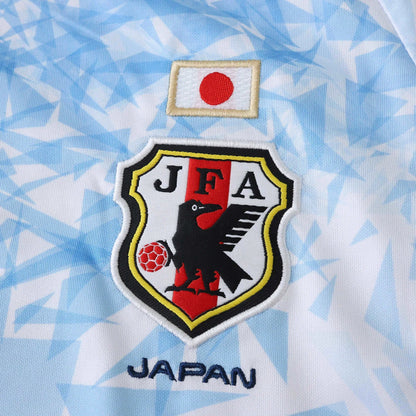 Camisa de futebol do Japão, vista de perto. Escudo JFA bordado com corvo e bola, bandeira japonesa e texto JAPAN em azul na camisa branca com detalhes em azul.