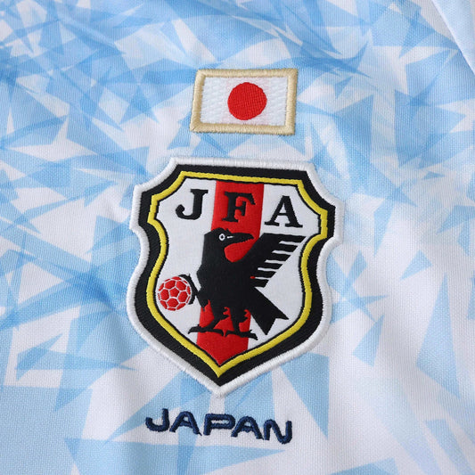 Camisa de futebol do Japão, vista de perto. Escudo JFA bordado com corvo e bola, bandeira japonesa e texto JAPAN em azul na camisa branca com detalhes em azul.
