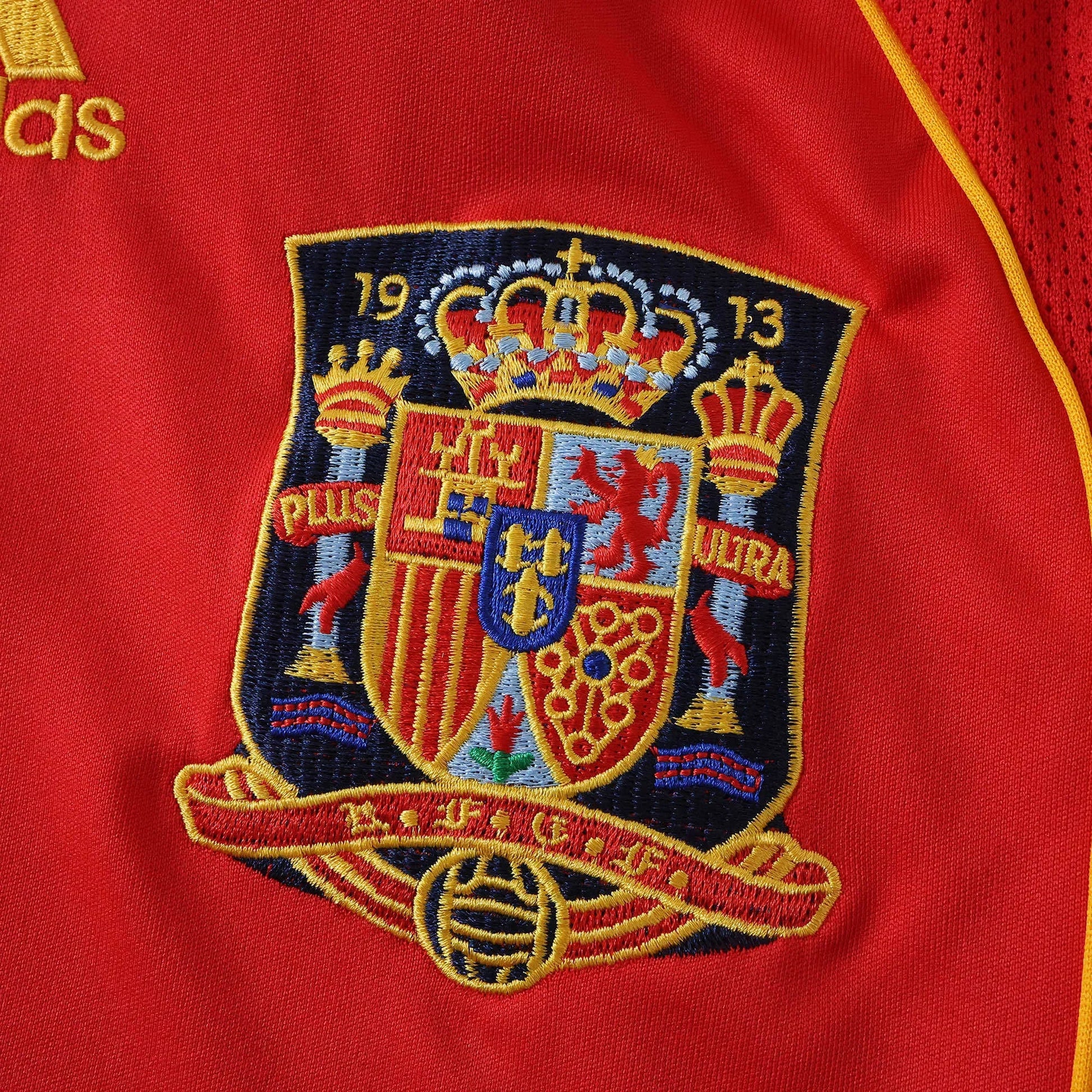 Close-up da camisa vermelha da Espanha, mostrando o escudo bordado em azul, dourado, vermelho e azul claro com coroas e as datas 19 e 13.