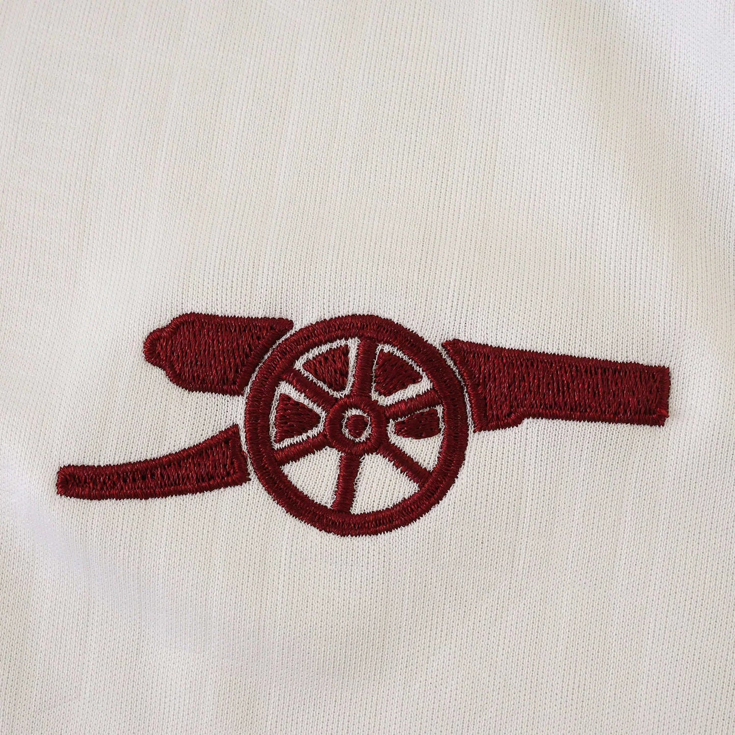 Detalhe do emblema bordado em vermelho-escuro de um canhão em tecido branco texturizado de camisa de futebol infantil.