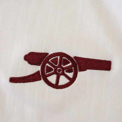 Detalhe do emblema bordado em vermelho-escuro de um canhão em tecido branco texturizado de camisa de futebol infantil.