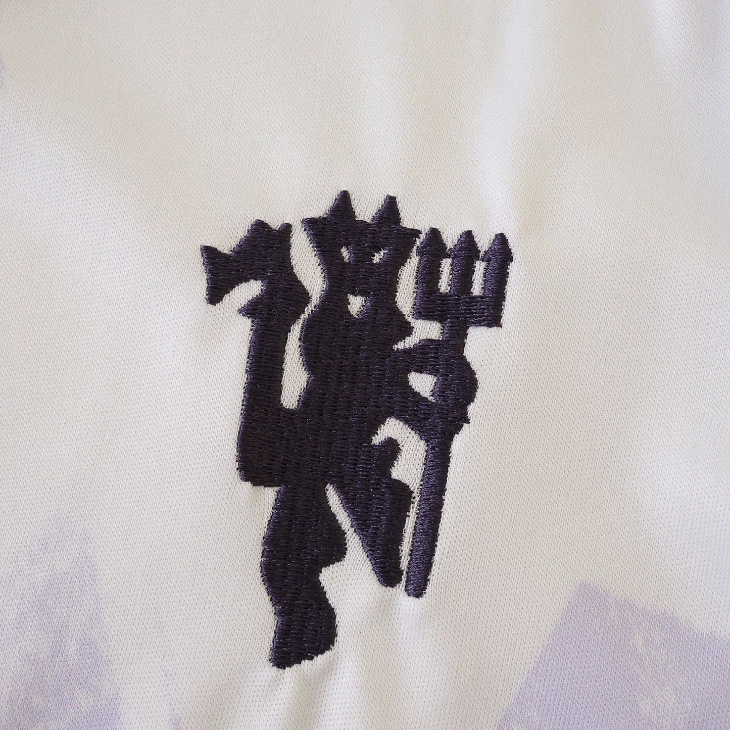 Detalhe do emblema bordado de um diabo roxo escuro segurando um tridente em uma camisa infantil branca do Manchester United com sutis padrões lilases.