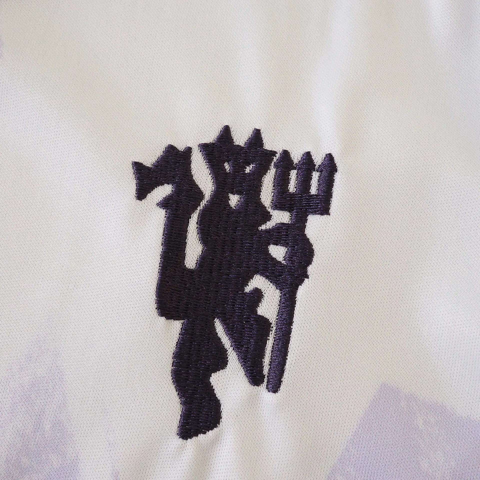 Detalhe do emblema bordado de um diabo roxo escuro segurando um tridente em uma camisa infantil branca do Manchester United com sutis padrões lilases.