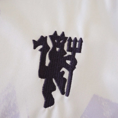Detalhe do emblema bordado de um diabo roxo escuro segurando um tridente em uma camisa infantil branca do Manchester United com sutis padrões lilases.