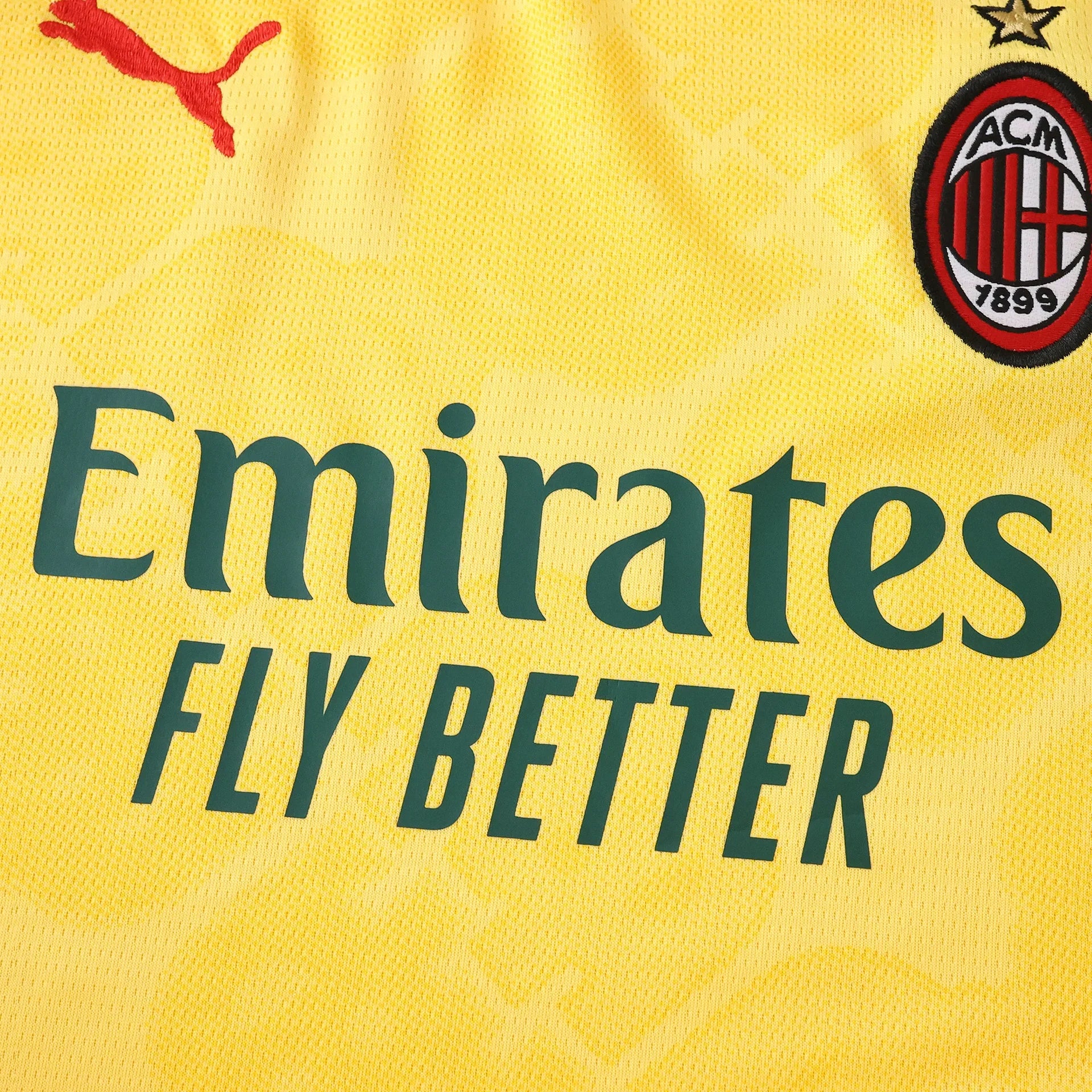 Close-up da camisa amarela do AC Milan infantil com escudo, logo PUMA vermelho e patrocinador Emirates FLY BETTER verde.