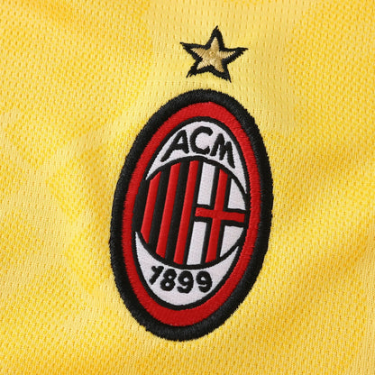 Close-up do escudo bordado do AC Milan e estrela dourada em camisa de futebol infantil amarela com textura de tecido.