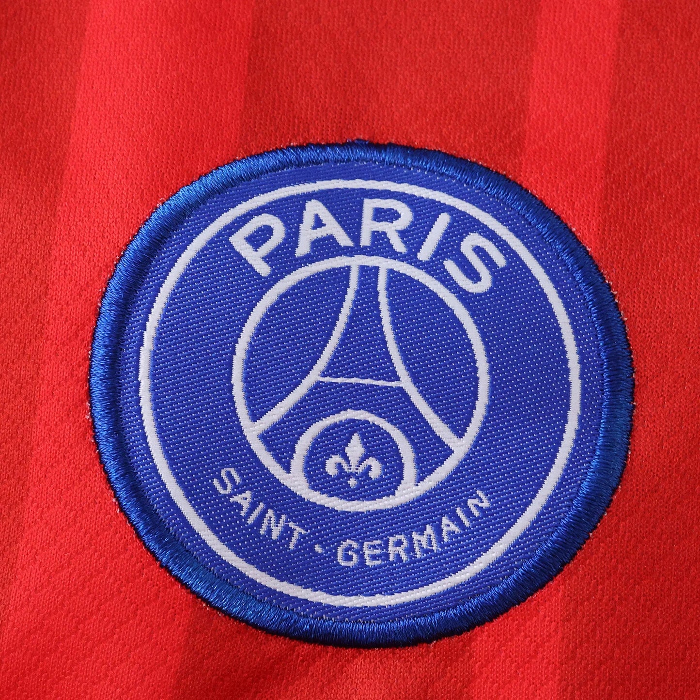 Detalhe do escudo bordado do Paris Saint-Germain em azul e branco, com a Torre Eiffel, na camisa de futebol infantil vermelha.