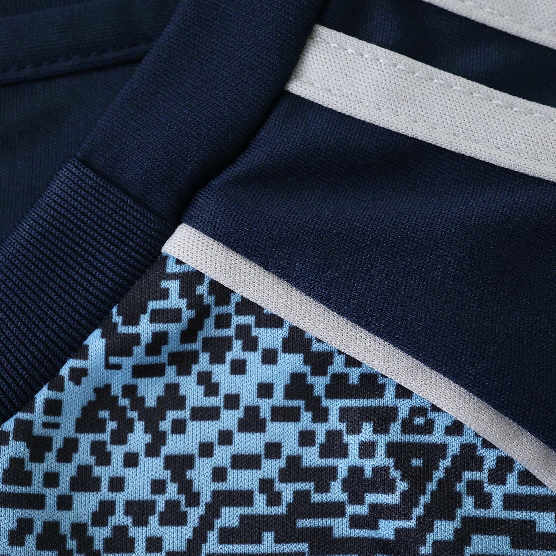 Close-up de kit de futebol infantil azul marinho com listras cinza claro e padrão pixelado em azul e preto.