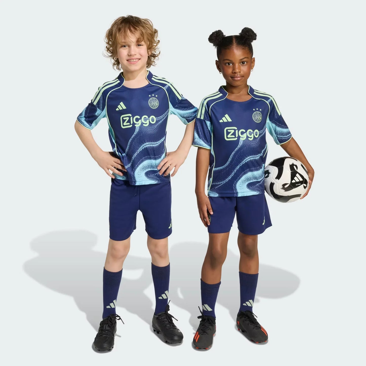 Duas crianças sorridentes vestindo o kit infantil do Ajax II em azul marinho com detalhes verde-limão. A menina segura uma bola de futebol.