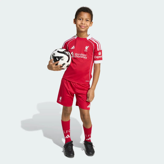 Menino sorridente veste kit de futebol infantil Liverpool FC 25/26 vermelho com shorts e meias, segurando bola de futebol.