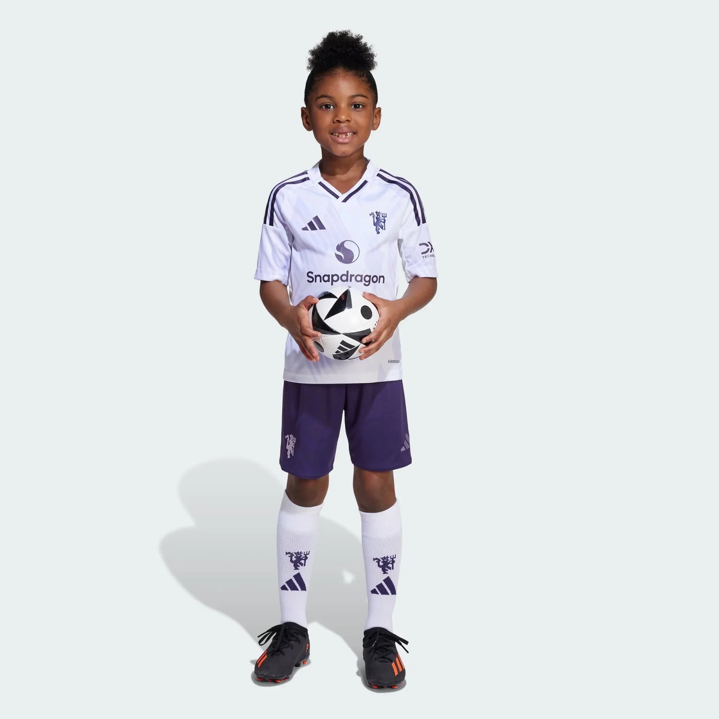 Criança sorridente veste kit infantil branco e roxo do Manchester United, segurando uma bola de futebol com as duas mãos.