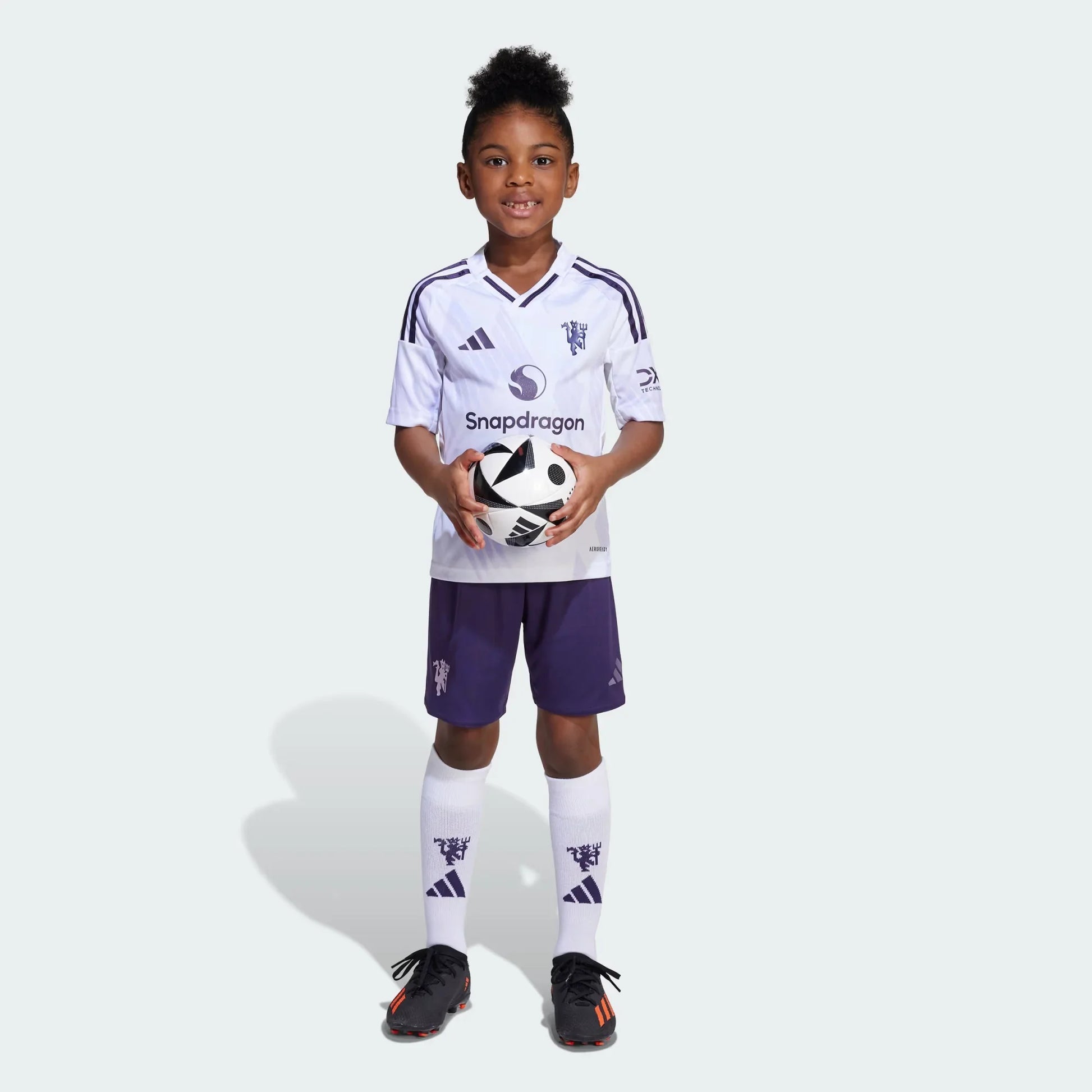 Criança sorridente veste kit infantil branco e roxo do Manchester United, segurando uma bola de futebol com as duas mãos.