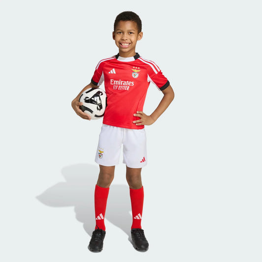 Menino sorridente em pé, vestindo kit de futebol vermelho e branco do Benfica, segurando uma bola preta e branca.