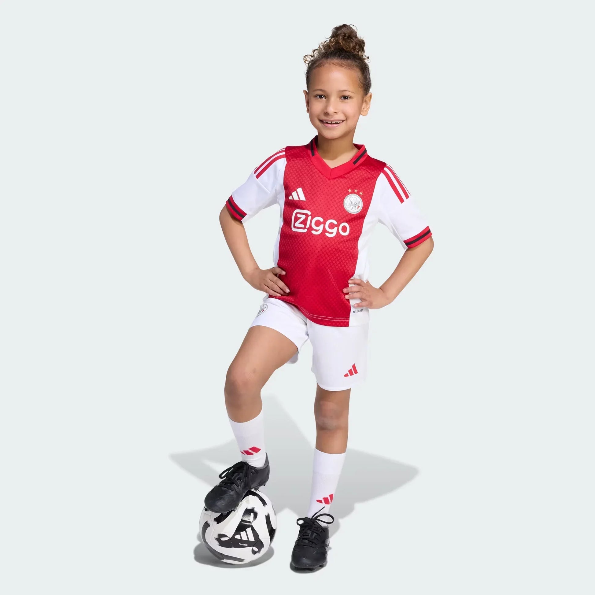 Criança sorridente veste kit do Ajax (camisa vermelha e branca, shorts brancos) com o pé na bola de futebol.