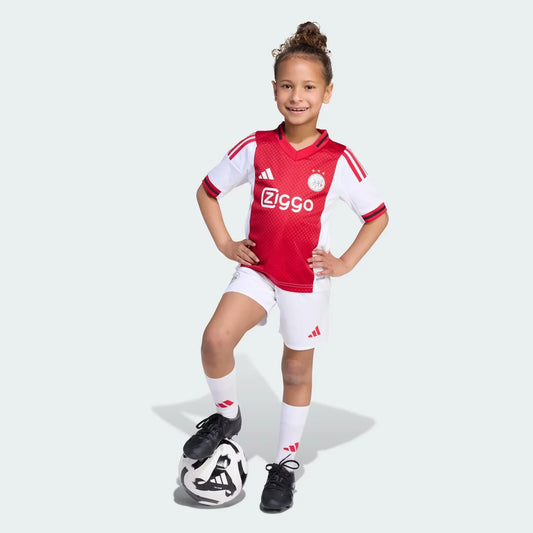 Criança sorridente veste kit do Ajax (camisa vermelha e branca, shorts brancos) com o pé na bola de futebol.