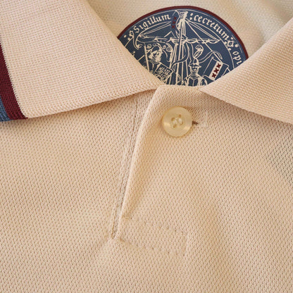 Detalhe da gola de camisa polo infantil creme com tecido piqué, botão claro, listras azul e bordô na gola. Emblema azul com dizeres e escudo XXX.