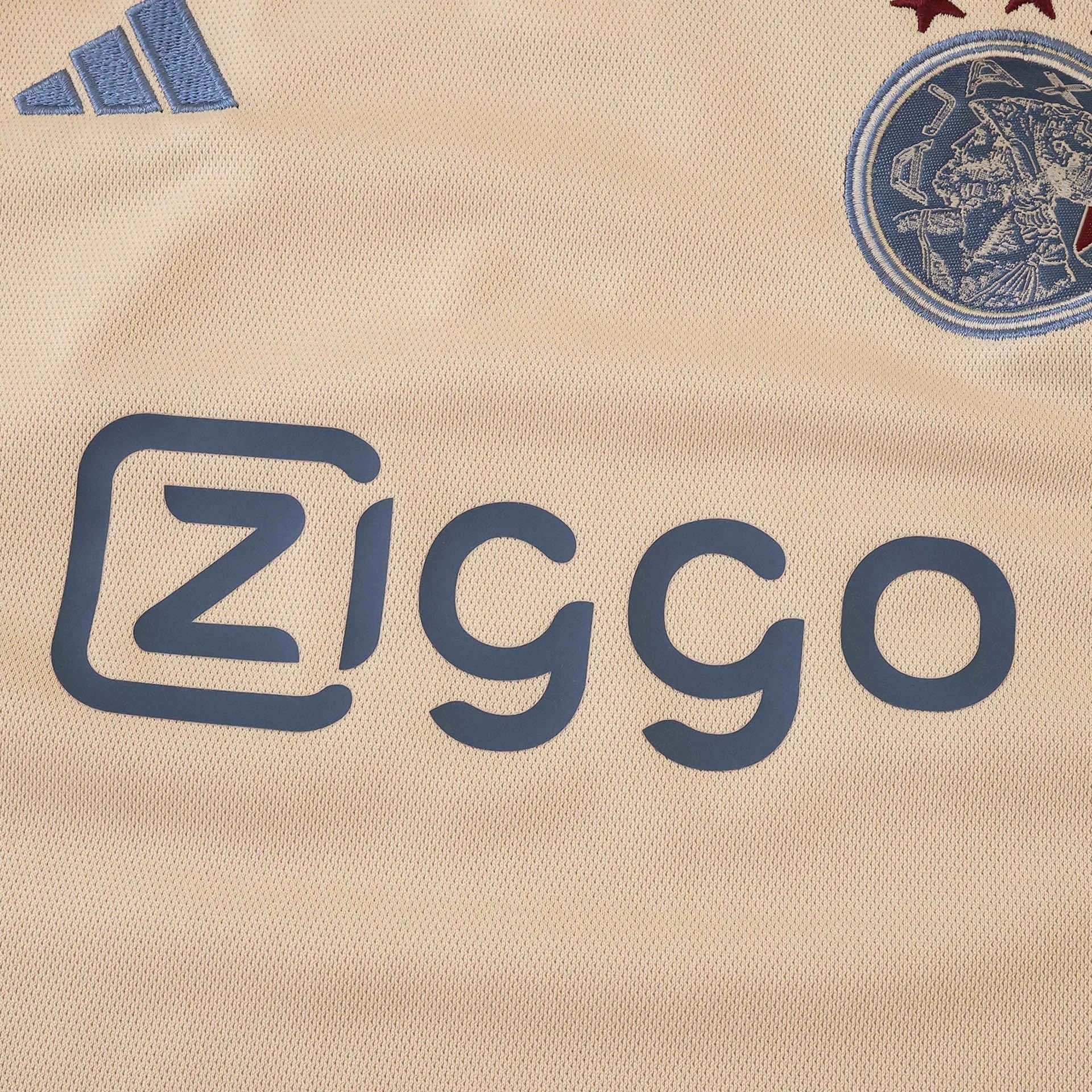 Detalhe da camisa creme claro do Ajax, exibindo o escudo azul com estrelas vermelhas, logo Adidas e patrocínio Ziggo azuis.