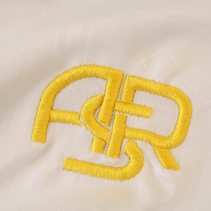Close-up de tecido branco com bordado dourado de letras entrelaçadas. Detalhe da textura do tecido e dos pontos do bordado.