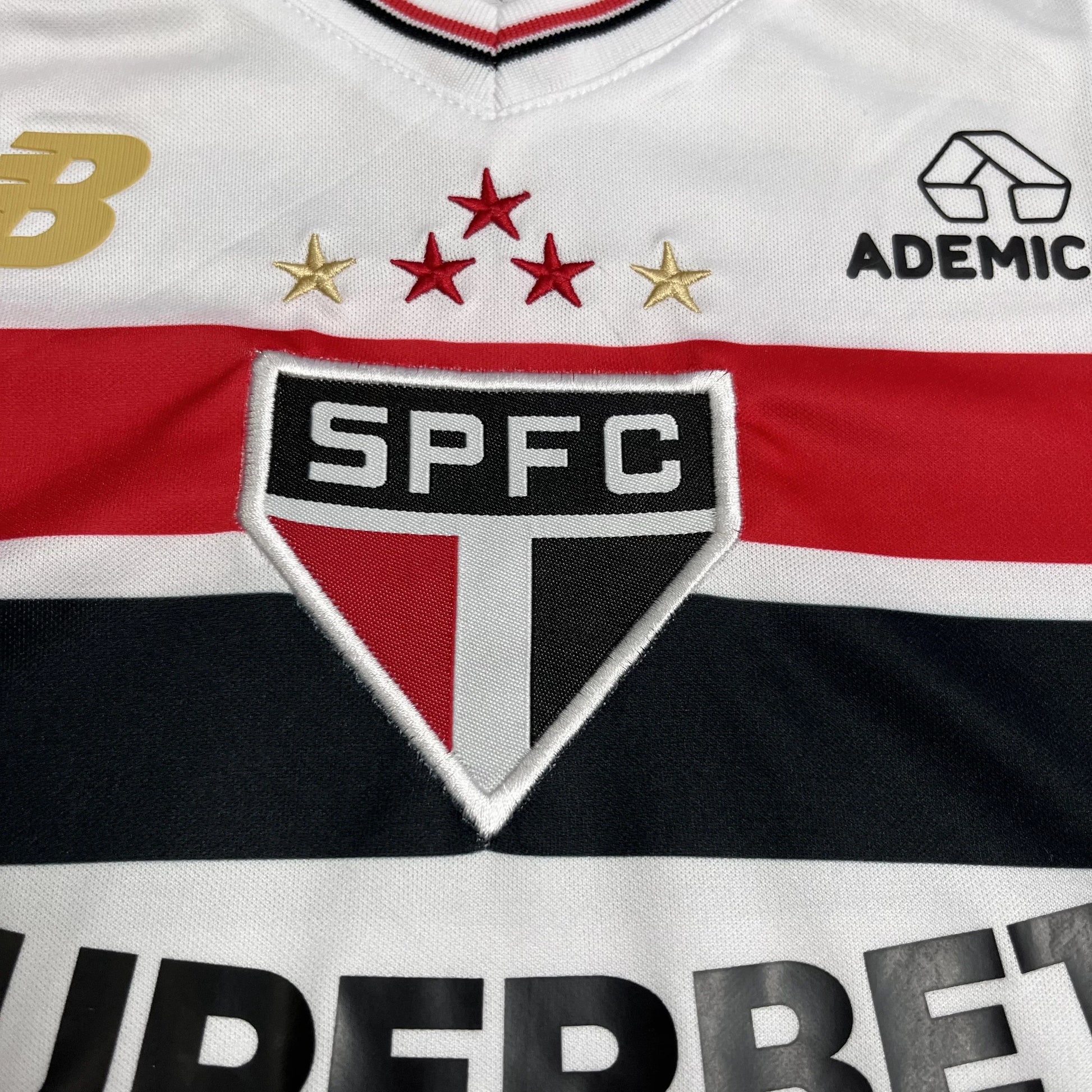 Close-up da camisa branca do São Paulo FC com escudo SPFC bordado, estrelas vermelhas e douradas, e listras horizontais vermelha e preta.