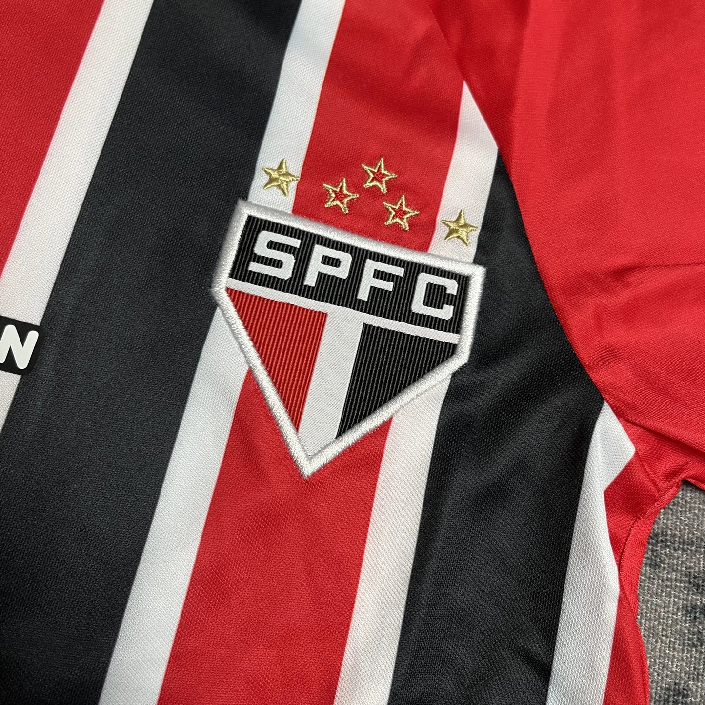 Close-up do escudo bordado SPFC com cinco estrelas douradas em camisa infantil com listras vermelhas, pretas e brancas.