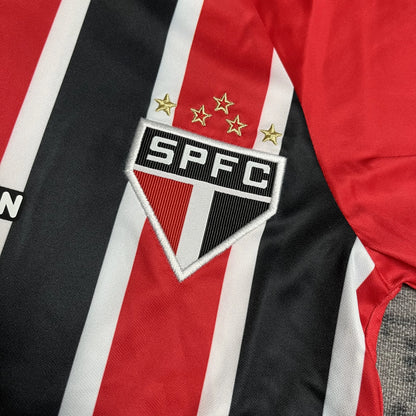 Close-up do escudo bordado SPFC com cinco estrelas douradas em camisa infantil com listras vermelhas, pretas e brancas.