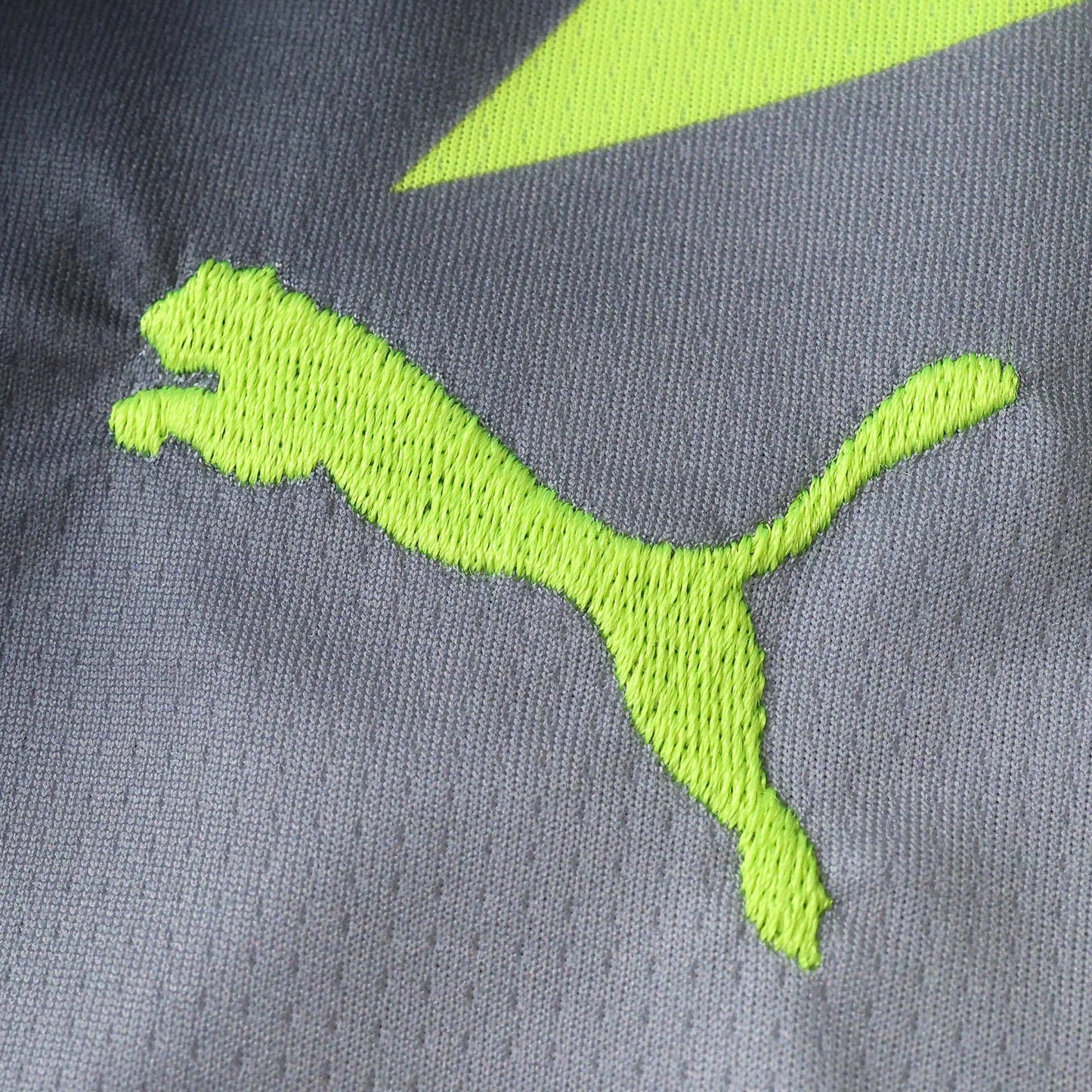 Close-up de tecido cinza com logo Puma bordado em verde neon e detalhe diagonal verde. Mostra a textura da malha esportiva.