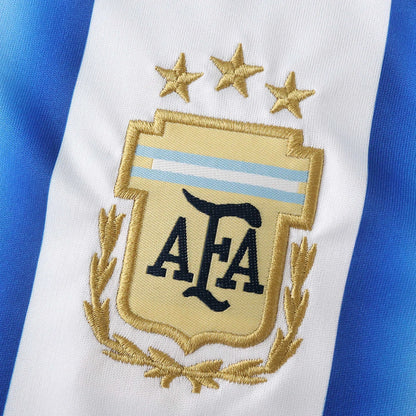 Detalhe do escudo bordado da AFA com três estrelas douradas sobre fundo listrado branco e azul.