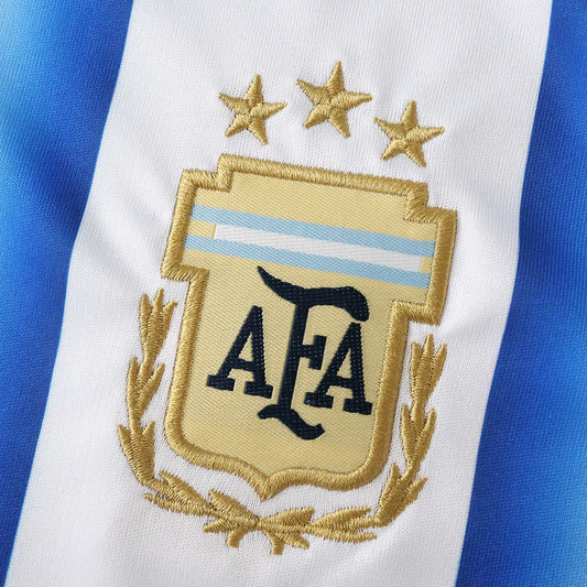 Detalhe do escudo bordado da AFA com três estrelas douradas sobre fundo listrado branco e azul.