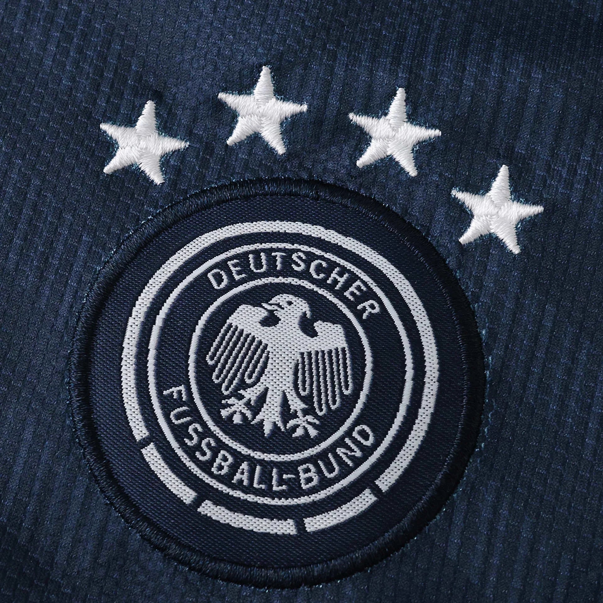 Detalhe do escudo bordado da seleção da Alemanha, com o brasão da DFB e a frase 'DEUTSCHER FUSSBALL-BUND'. Quatro estrelas brancas de campeão mundial estão acima do escudo, tudo em tecido azul marinho.