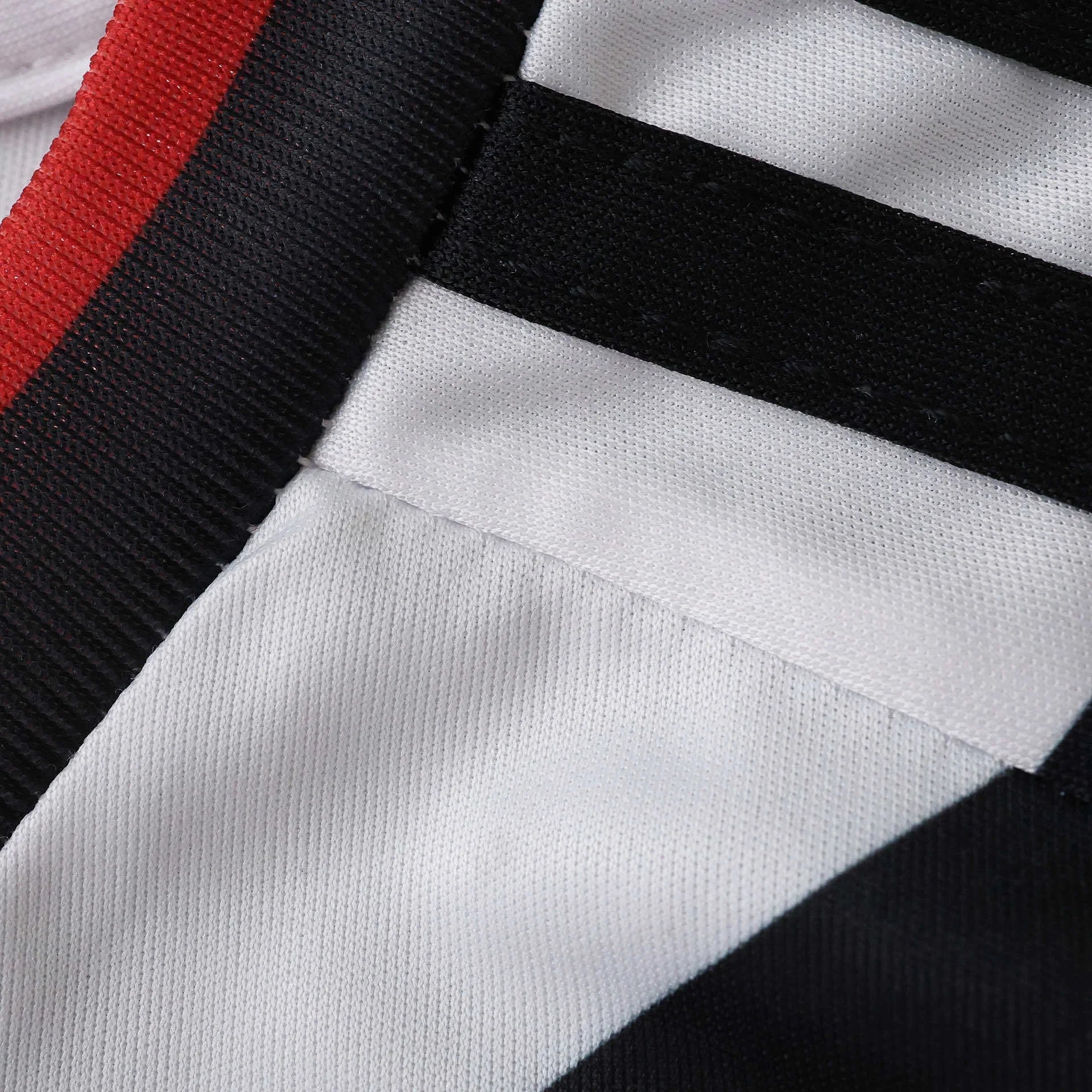 Close-up de tecido branco com listras pretas diagonais, e faixa vermelha e preta na borda superior. Textura do tecido visível.