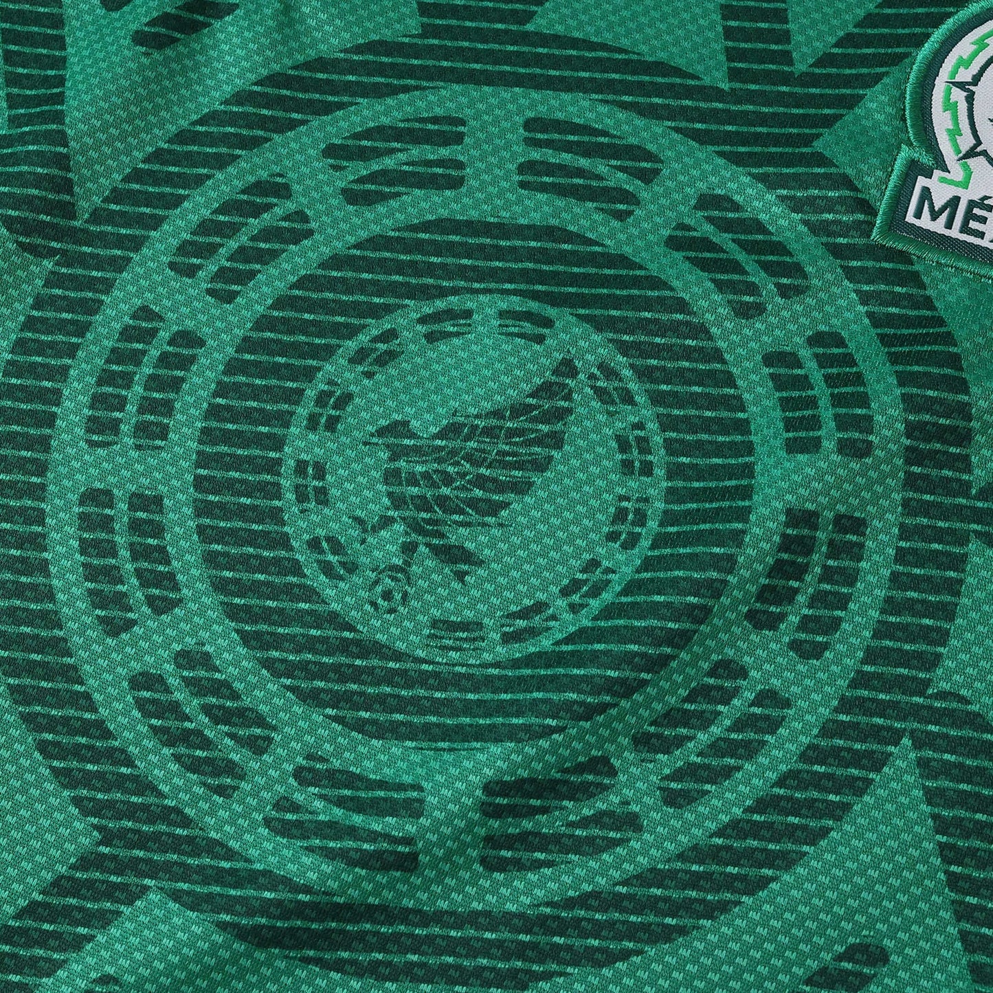 Camisa de futebol infantil do México verde escura com padrão circular sutil do calendário asteca e brasão parcial da seleção.