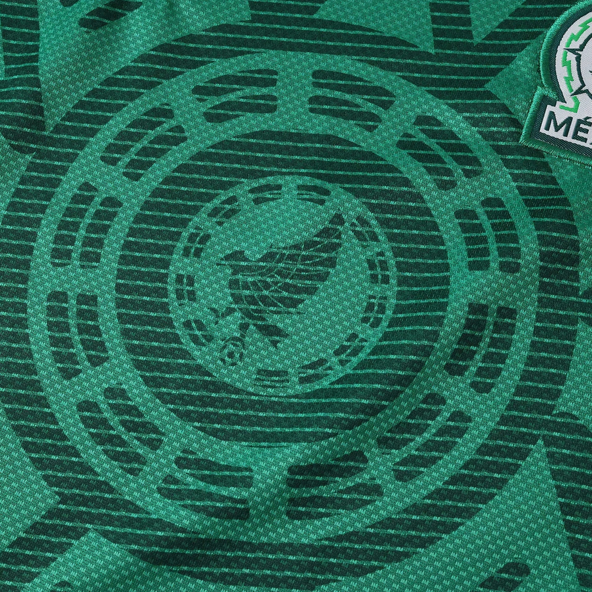 Camisa de futebol infantil do México verde escura com padrão circular sutil do calendário asteca e brasão parcial da seleção.