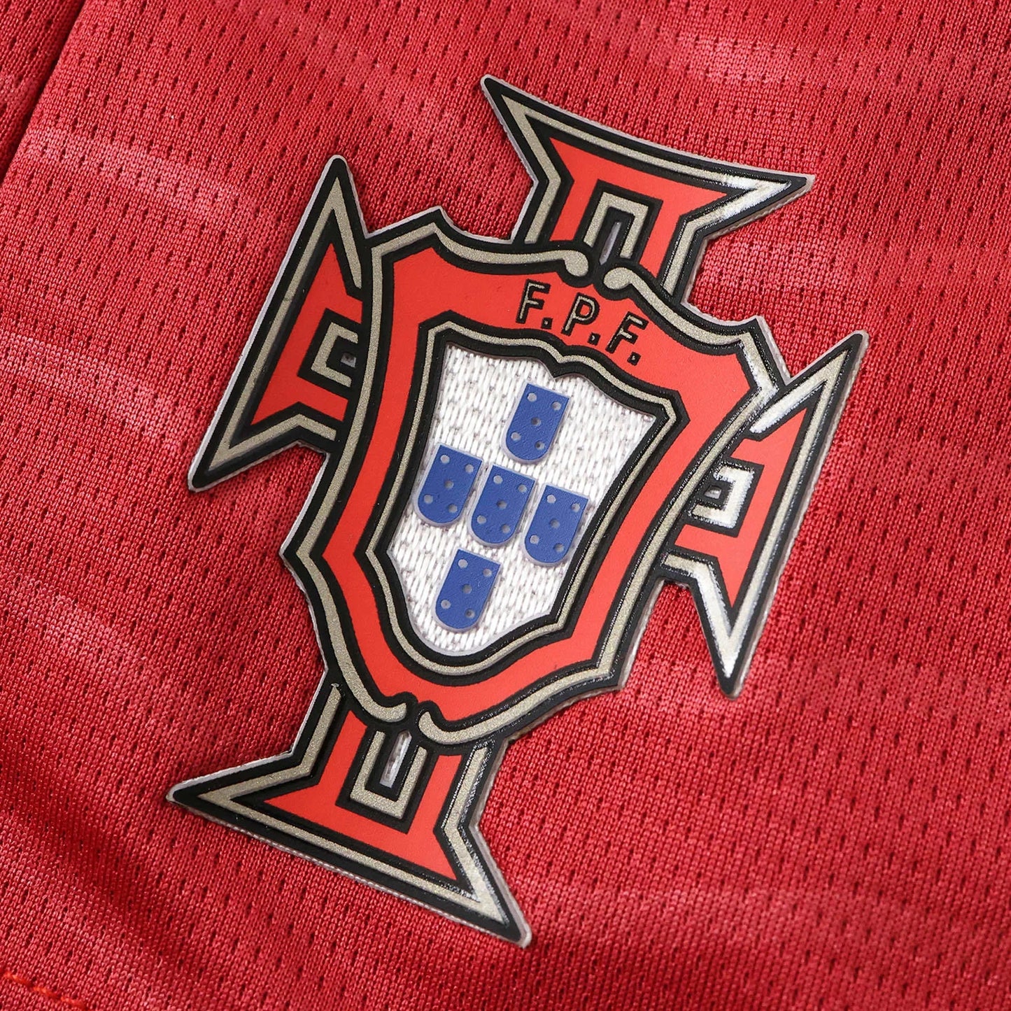 Close-up do escudo da seleção de Portugal em uma camisa de futebol infantil vermelha, mostrando o escudo F.P.F. com detalhes azuis e brancos.