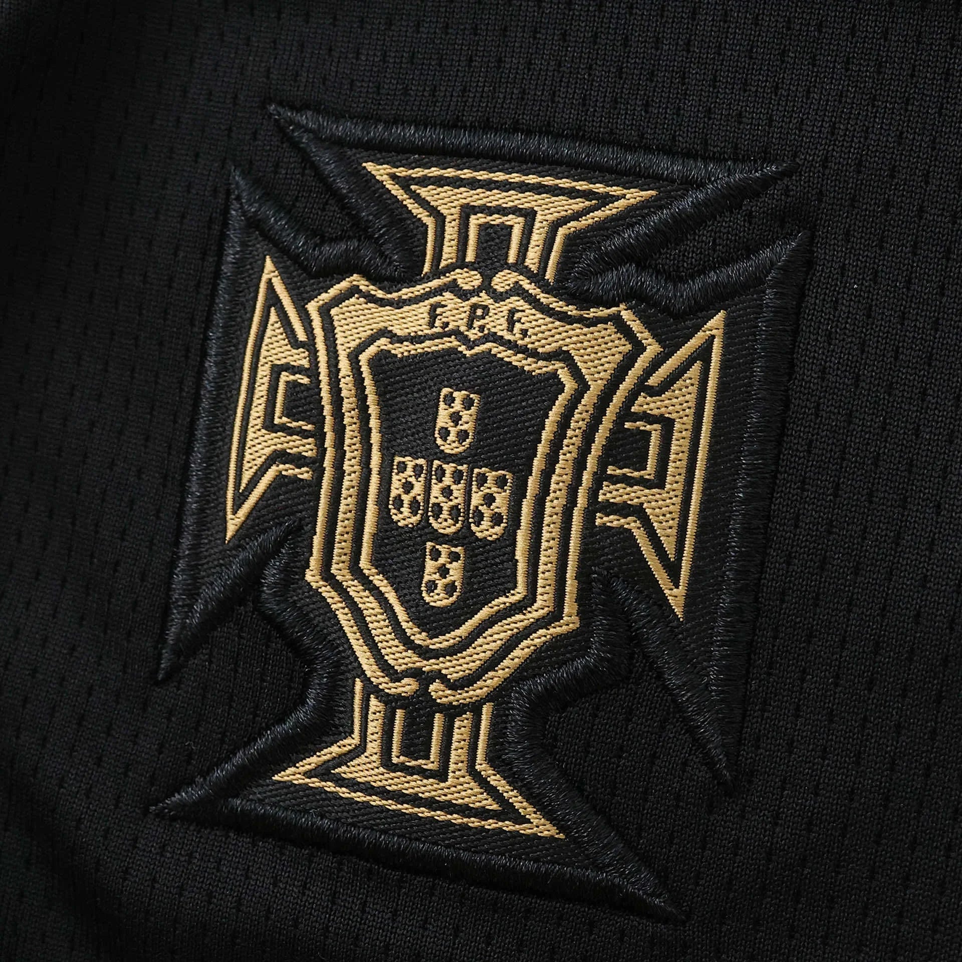 Emblema bordado da seleção de Portugal em dourado sobre tecido preto de malha, exibindo a Cruz de Cristo e escudo com FPF.