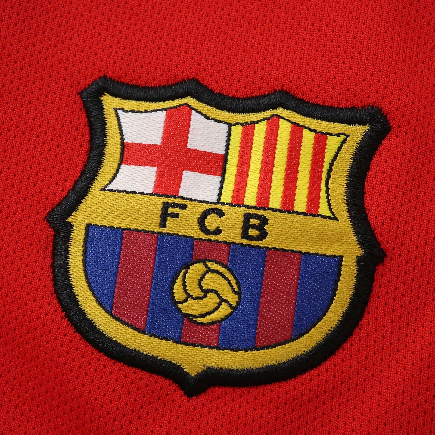 Close-up do escudo bordado do FC Barcelona em uma camisa de tecido vermelho texturizado.