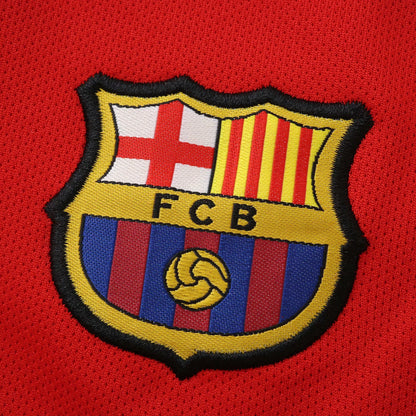 Close-up do escudo bordado do FC Barcelona em uma camisa de tecido vermelho texturizado.