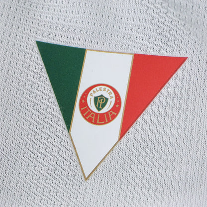 Close-up de um emblema triangular com as cores da bandeira italiana (verde, branco, vermelho) e o logo 'PALESTRA ITALIA' em uma camisa branca com textura.