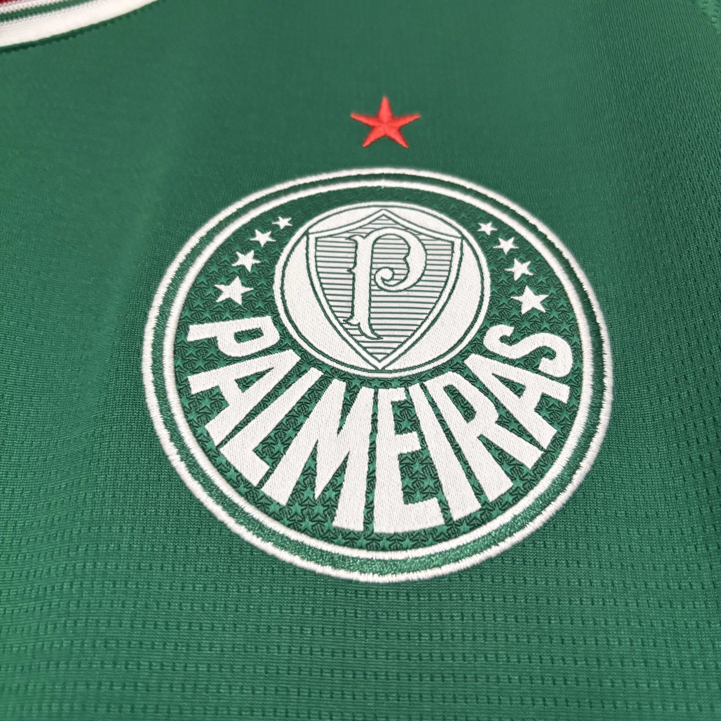 Close detalhado do escudo oficial do Palmeiras bordado em branco sobre o tecido verde da camisa, apresentando a estrela vermelha acima do emblema.