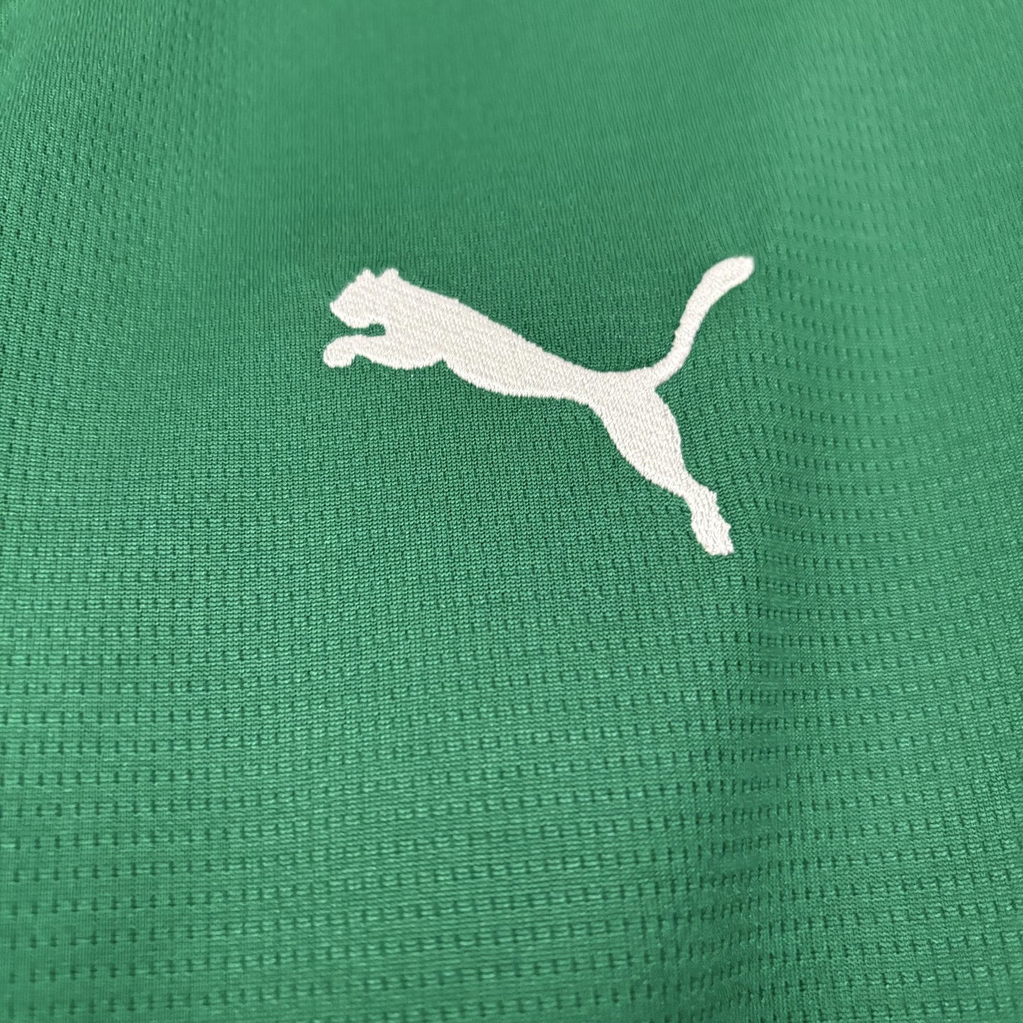 Close macro da textura do tecido tecnológico da camisa na cor verde, evidenciando o logo da Puma bordado em branco.