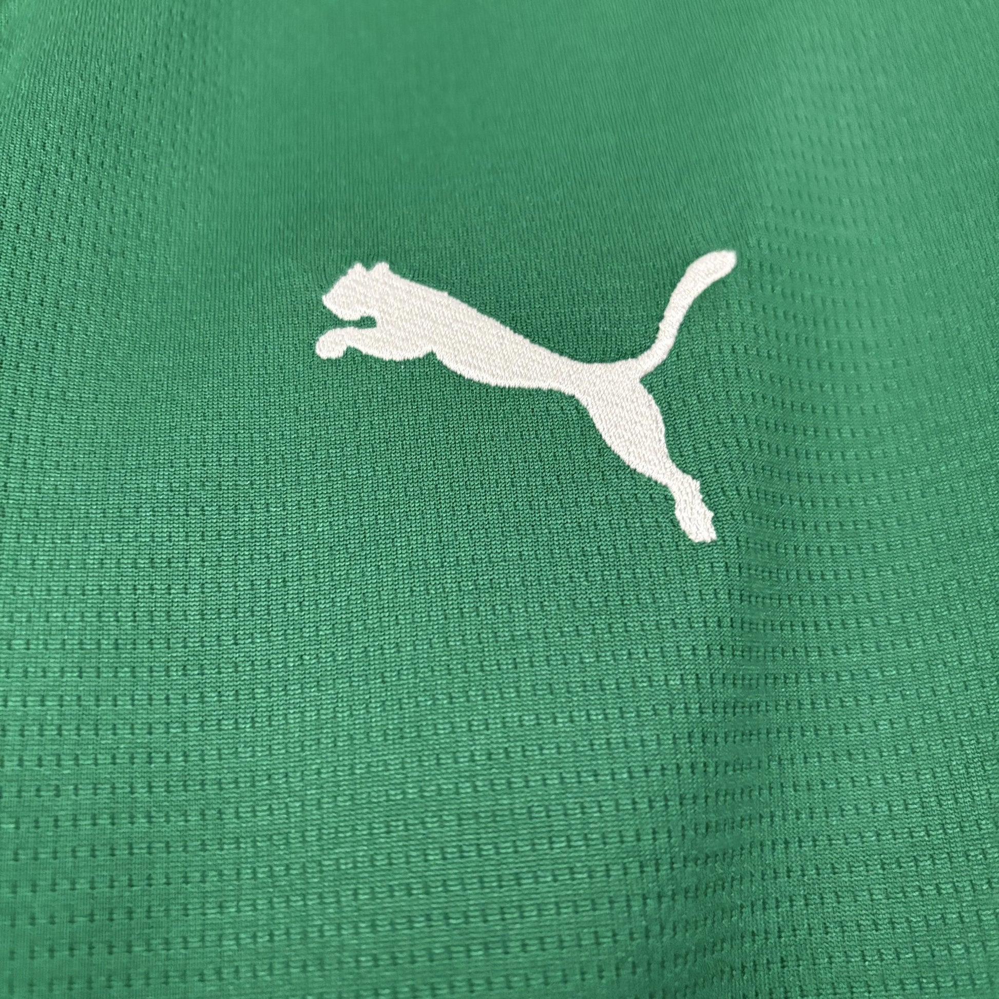 Close macro da textura do tecido tecnológico da camisa na cor verde, evidenciando o logo da Puma bordado em branco.