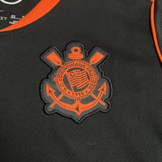 Close detalhado do escudo do Corinthians emborrachado em tons de laranja e preto, aplicado sobre o tecido tecnológico preto da camisa.