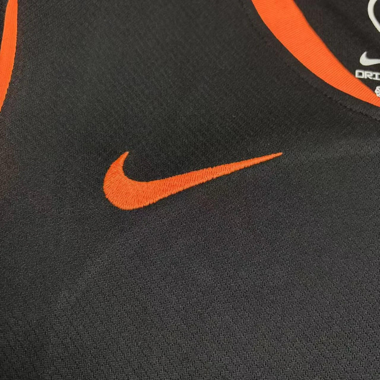 Close do logotipo "Swoosh" da Nike bordado em laranja no lado direito do peito, contrastando com o fundo preto da camisa.