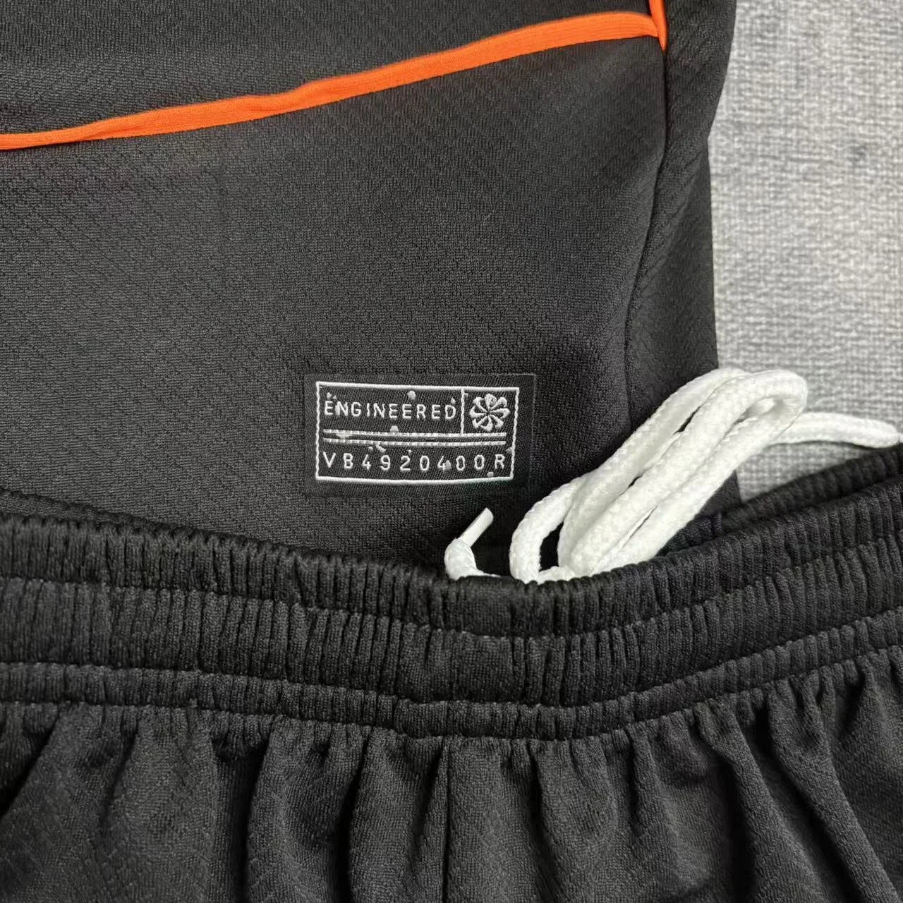 Close da etiqueta de autenticidade prateada aplicada na barra da camisa e do detalhe do cordão de ajuste branco no cós elástico do calção preto.