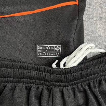 Close da etiqueta de autenticidade prateada aplicada na barra da camisa e do detalhe do cordão de ajuste branco no cós elástico do calção preto.
