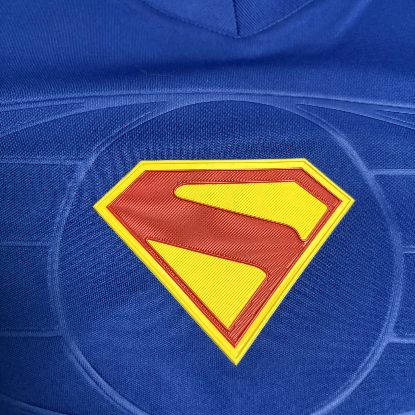 Close detalhado do icônico escudo do Super-Homem (um "S" vermelho sobre fundo amarelo em formato de diamante) aplicado ao centro da camisa azul.